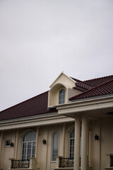 Obraz premium Low-angle view of a classical building featuring a red tiled roof, arched dormer window, and elegant architectural details against an overcast sky, highlighting timeless design and formal architecture