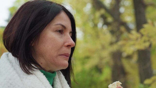 woman sipping from mug outdoors wrapped in white blanket and green sweater, solitary hiker pausing for tea and bread during autumn picnic in forest, closeup portrait showing contemplative expression