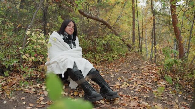 sitting woman wrapped in blanket forest, leather boots and shawl on leafstrewn path, soft overcast light, pensive expression, quiet solitude, textured blanket, fall foliage, rustic trees, meditative
