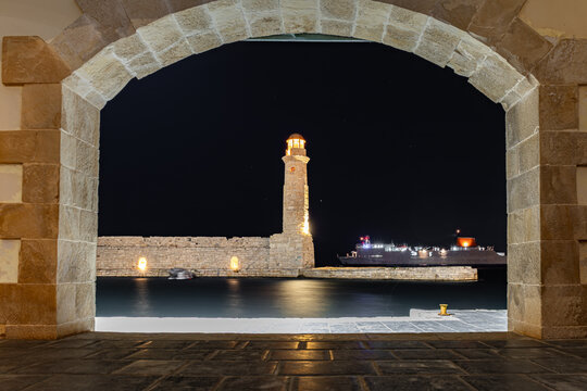 View of an illuminated lighthouse rising against the inky night sky, framed by a stone archway with shimmering water, Rethimno, Rethymno, Greece.
