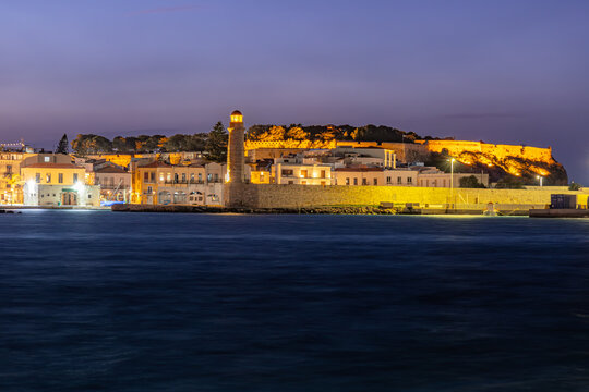 View of a glowing lighthouse stands tall against the twilight sky, illuminating the ancient fortress and waterfront buildings, Rethimno, Rethymno, Greece.
