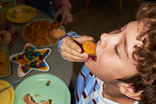 Caucasian boy eating chicken nugget at table, tilting head back with mouth open, surrounded by plates of snacks and colorful candies, enjoying food during meal or party