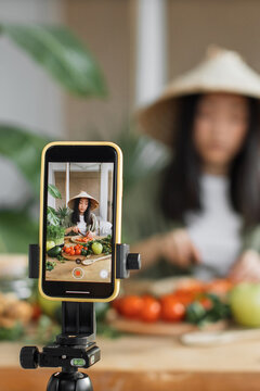 A young Asian woman wearing a conical hat films herself preparing fresh vegetables for a cooking tutorial on her smartphone