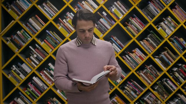 Slowmo portrait of young Caucasian man taking book from library shelf with modern diagonal design, reading it and then smiling at camera