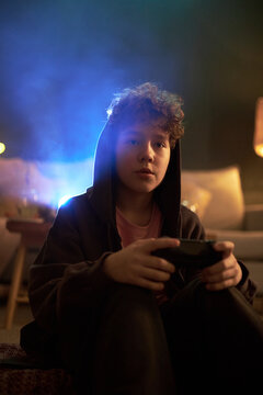 Portrait of Caucasian teenage boy sitting indoors holding game controller wearing hoodie looking into camera illuminated by blue light background showing focused expression