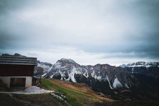 View of a rustic wooden chalet perched on a vibrant green hillside, overlooking the majestic, snow-dusted Dolomites under a moody sky, Urtijei, Trentino-South Tyrol, Italy.