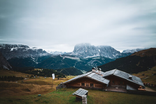 View of a rustic wooden chalet nestled in the grassy highlands beneath the towering, snow-dusted Dolomites under a cloudy sky, Urtijei, Trentino-South Tyrol, Italy.