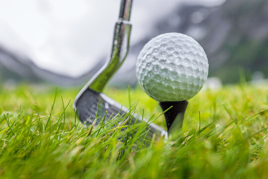Golf club and ball on wet green field with mountains in background.