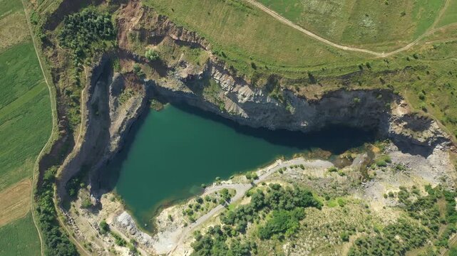 Aerial view of the stunning Racos Quarry Lake, where blue waters meet rocky terrain, contrasted by surrounding green fields, Racos, Judetul Brasov, Romania.
