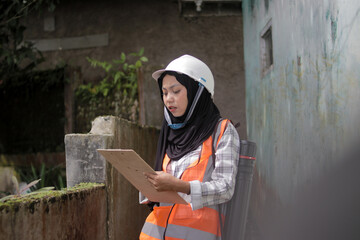 Young Asian Muslim woman working as a professional civil engineer or site inspector wears a hijab, white hard hat, and orange safety vest over a plaid shirt. reviewing documents at construction site