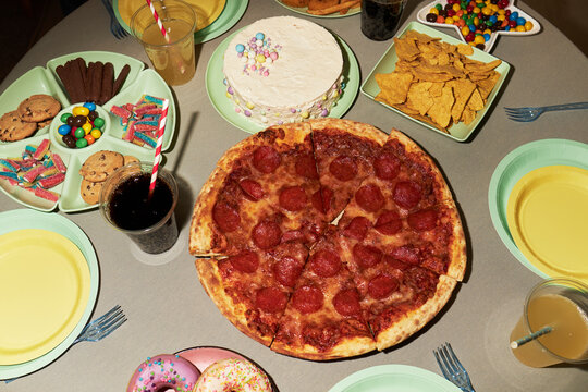 Overhead view showing pepperoni pizza, cake, cookies, donuts, tortilla chips, candy, and soft drinks arranged on table with empty plates and cups prepared for group gathering