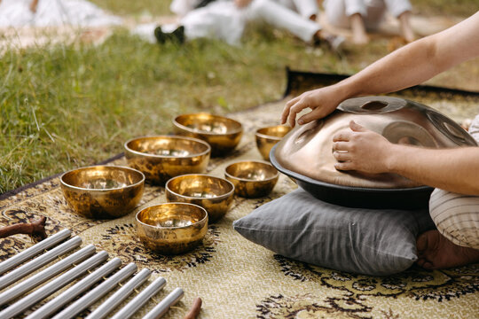 Person playing a handpan surrounded by singing bowls during an outdoor sound healing ceremony in nature.