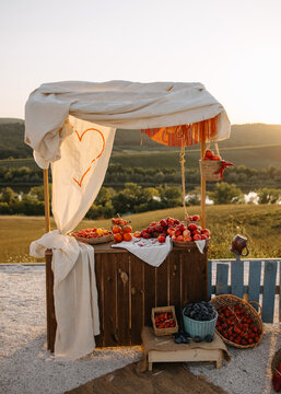 Rustic outdoor fruit market stand at sunset with apples, tomatoes, and grapes, styled for a countryside wedding or event. Romantic farm aesthetic with natural light.