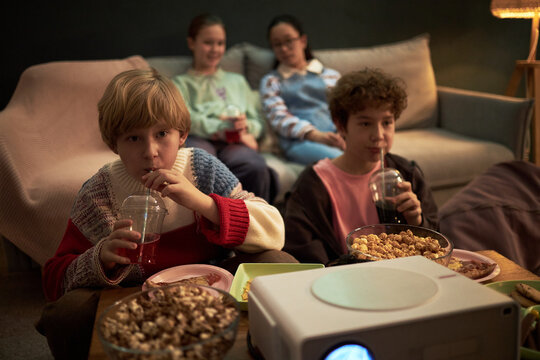 Two Caucasian kids sitting in foreground drinking from plastic cups and eating popcorn while watching movie on projector, two girls sitting on sofa in background watching screen