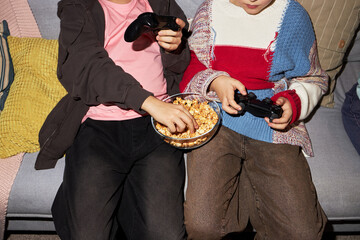 Two children sitting on sofa holding video game controllers and sharing bowl of popcorn, one child reaching for snack while both focusing on playing video game together © DragonImages