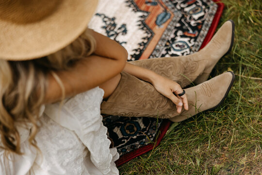 Woman wearing suede western boots sitting on a patterned rug outdoors, relaxed summer lifestyle scene with natural textures, grass, and boho-inspired fashion details.