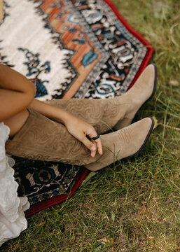 Boho dressed woman wearing beige suede cowboy boots while sitting on a patterned rug outdoors. Rustic detail with western style, natural light, and relaxed countryside vibes.