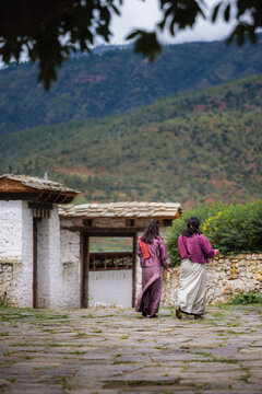 View of two women in traditional dresses stroll across a stone courtyard towards an ancient gateway, framed by mountains, Rinpung Dzong, Paro, Bhutan.