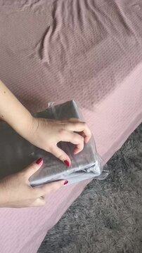 A woman's hands open a package with a new sheet with an elastic band against the background of the bed. New grey cotton bedding