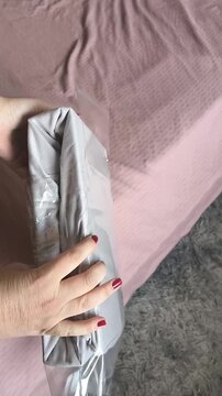 A woman's hands open a package with a new sheet with an elastic band against the background of the bed. New grey cotton bedding
