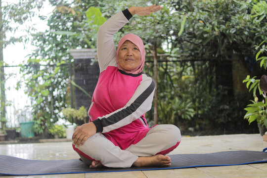Middle-aged Asian woman wearing a hijab performs yoga exercises on her home terrace. Home fitness, wellness, and healthy lifestyle concept.