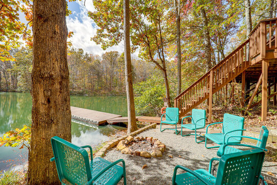 View of turquoise chairs encircle a stone-ringed fire pit near a tranquil lake reflecting the vibrant autumn foliage in Hamden, Ohio, United States.