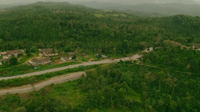 Aerial view of houses nestled amidst dense, verdant forests, dissected by winding roads with sparse traffic, Sakleshpura, Karnataka, India.