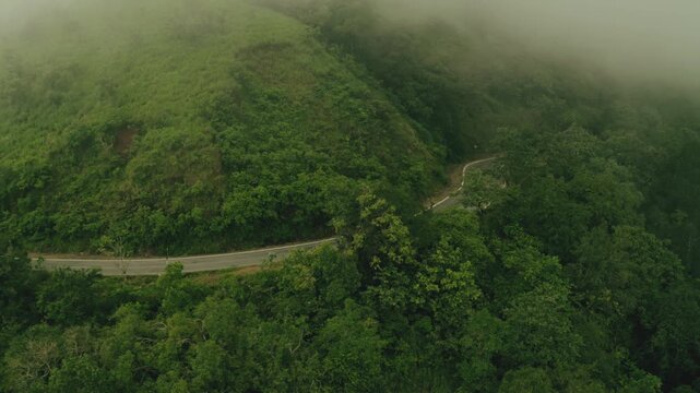 Aerial view of a grey road winding through verdant hills and dense green trees, shrouded in mist, creating a serene landscape, Sakleshpura, Karnataka, India.