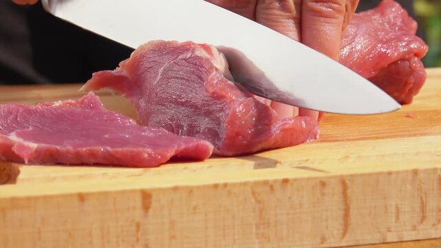 Knife cutting fresh raw pork meat into small pieces on a wooden board. Cooking process in the kitchen. Preparation of ingredients for meal.