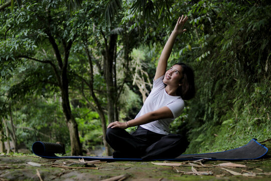Young Asian woman with short hair wearing a white shirt performs yoga exercises on a black mat in a forest setting. Mindfulness, wellness, and healthy lifestyle concept.