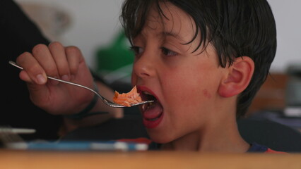 Young boy eats salmon from fork held by adult, mid-bite moment of home dining with visible parent...