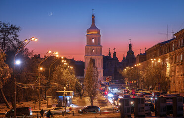 The waxing crescent moon sets behind the bell tower of St. Sophia Cathedral. Twilight colors the sky as city lights glow, revealing a winter view of Sophia Square in Kyiv.