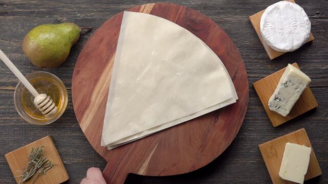 Wooden board with phyllo dough sheets placed on table with pear and cheese pie ingredients. Top view of baking preparation process in kitchen