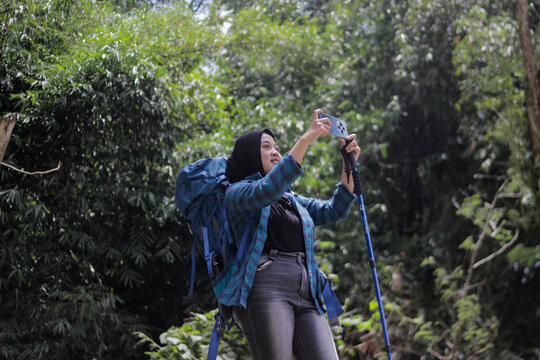 Young Asian woman wearing a hijab and flannel shirt carries a backpack and trekking pole while taking a landscape photo with her phone during outdoor hike, hiking, and outdoor adventure concept