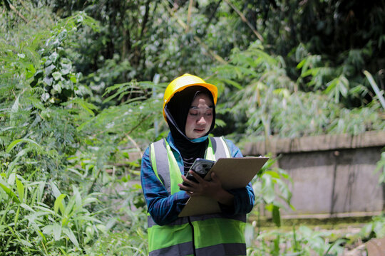 Asian female eco volunteer wearing hijab and hard hat holds a clipboard while inspecting trees in a nature reserve. Forestry engineering, environmental supervision, and conservation fieldwork concept