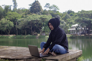 Fototapeta premium Young Asian woman wearing a hijab works on a laptop beside a lake in an outdoor setting. Remote work lifestyle, digital nomad, freelance work, online business, studying, and flexible workspace concept