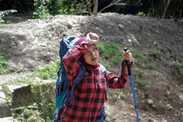 Tired young Asian woman hiker wearing a hijab and red flannel shirt, carrying a mountain backpack and holding a trekking pole while wiping sweat from her forehead during an outdoor hike