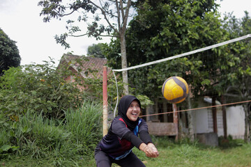 Young Asian woman wearing a hijab practices passing a volleyball on an outdoor volleyball court in a rural village setting. Grassroots sports, casual physical activity, and an active lifestyle concept