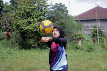 Young Asian woman wearing a hijab practices passing a volleyball on an outdoor volleyball court in a rural village setting. Grassroots sports, casual physical activity, and an active lifestyle concept