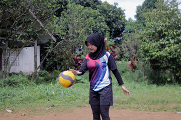 Young Asian woman volleyball player wearing a hijab practices serving on an outdoor volleyball court in a rural village setting. Sports training, grassroots athletics, and active lifestyle concept