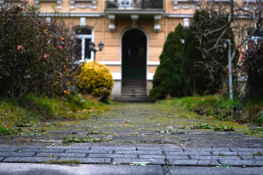 Low angle focus on an old stone paved walkway leading to the blurred arched entrance of a classic vintage residential building surrounded by garden bushes.
