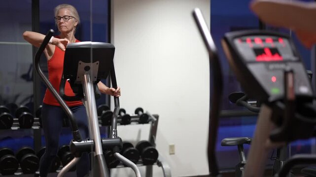 Mature woman uses elliptical machine for fitness in gym during evening hours with weights in background and focused expression.