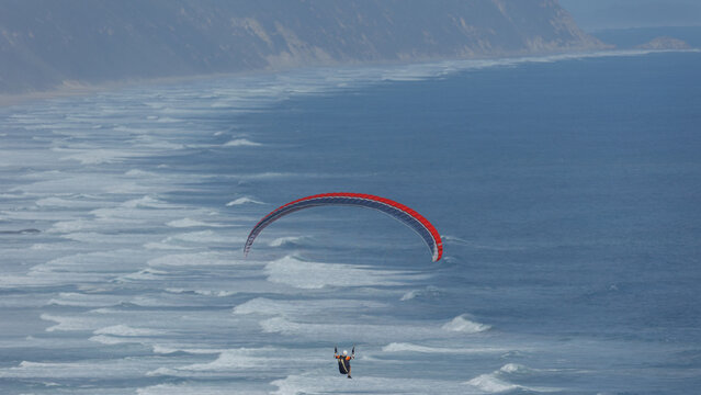 View of a paraglider soars against a backdrop of ocean waves meeting sandy shores, creating a dynamic aerial dance, Wilderness, Western Cape, South Africa.