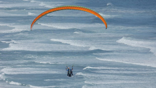 View of a paraglider soars above the turbulent, white-tipped waves crashing against the shore, a dance of freedom against the ocean's raw power, Wilderness, Western Cape, South Africa.