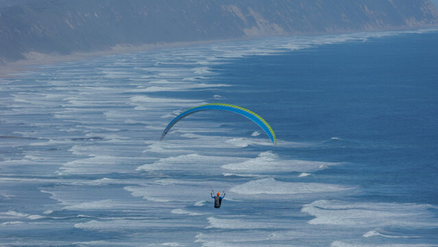 View of paraglider soars above the foamy turquoise waves meeting the sandy shore under a hazy sky, a dance of freedom and nature's beauty, Wilderness, Western Cape, South Africa.