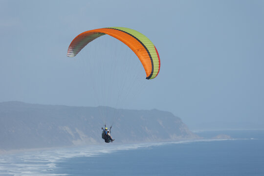 View of a paraglider soars above the rugged cliffs and foamy waves, a symphony of orange, green, and blue against the hazy sky, Wilderness, Western Cape, South Africa.