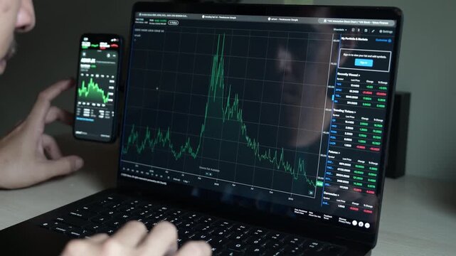 close up smartphone with laptop and open stock exchange, trading Online app interface. Man hand scrolling touch screen analysing chart forex crypto currency.