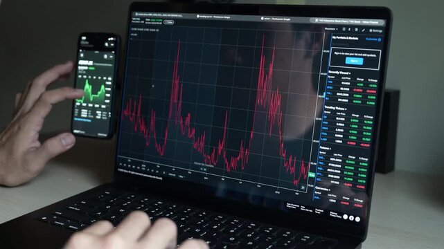 close up smartphone with laptop and open stock exchange, trading Online app interface. Man hand scrolling touch screen analysing chart forex crypto currency.