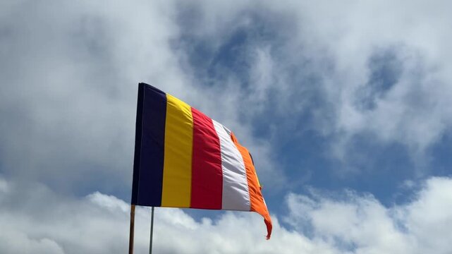 Buddhist flag waves on a tall pole against a cloudy sky, its bright stripes moving in the wind. Buddhist tradition.
