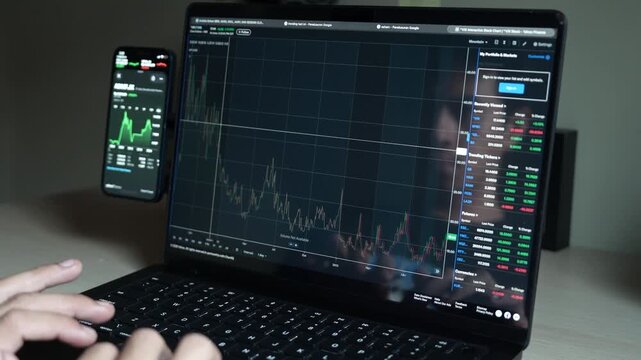 close up smartphone with laptop and open stock exchange, trading Online app interface. Man hand scrolling touch screen analysing chart forex crypto currency.